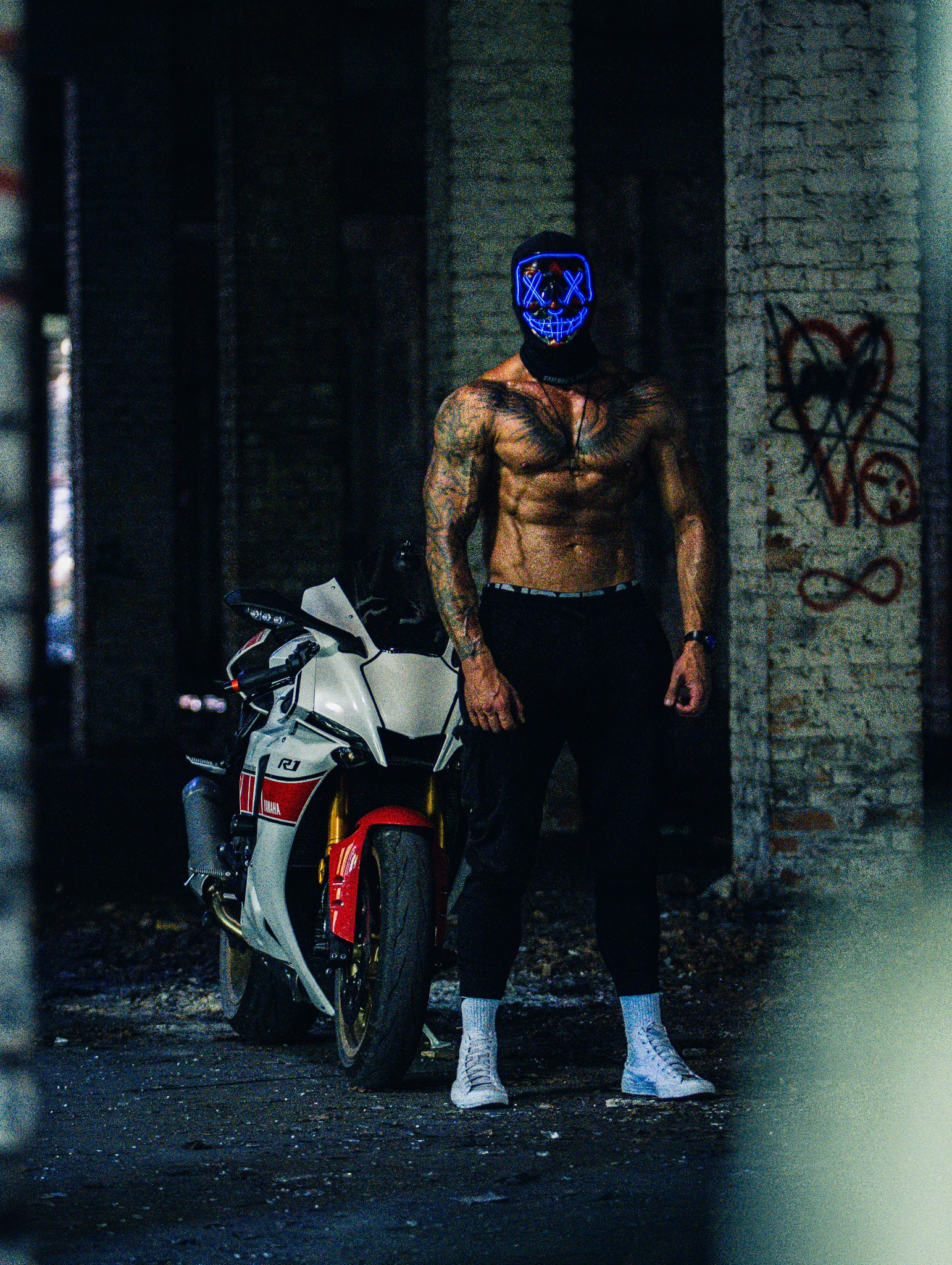 Mężczyzna w świecącej masce stoi obok motocykla Yamaha R1 w opuszczonym budynku. Tatuaże na ciele, ciemne spodnie i białe buty. Graffiti na ceglanej ścianie w tle.
