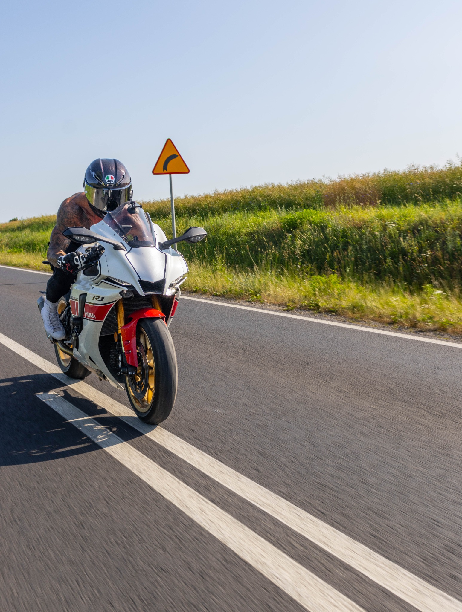 Biker w kasku AGV na białym motocyklu Yamaha R1, dynamicznie pokonujący asfaltową drogę z żółtym znakiem ostrzegawczym zakrętu w tle.