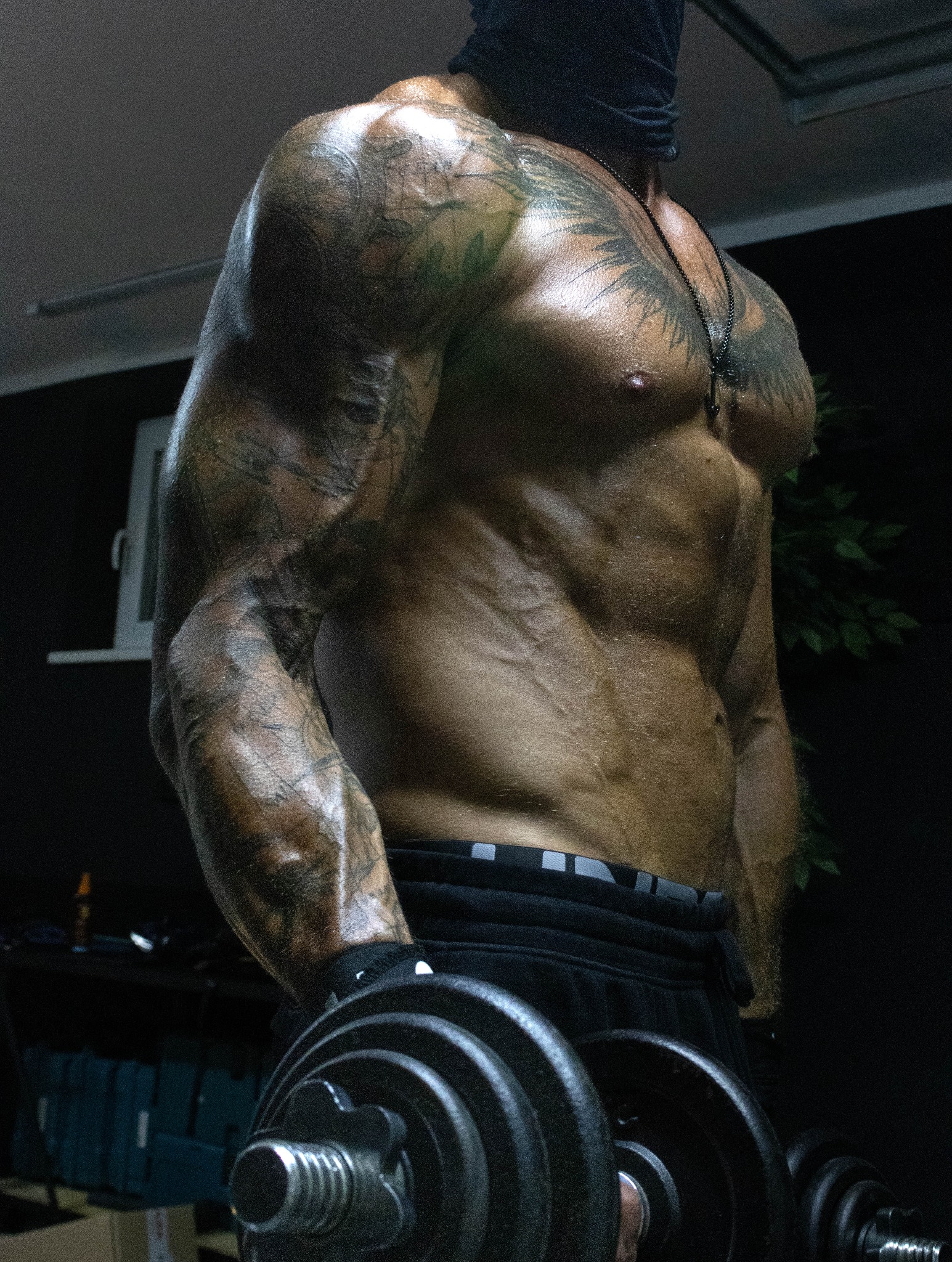 Mężczyzna z tatuażami, w czarnej masce, ćwiczy z hantlami. Widoczne napięte mięśnie klatki piersiowej i ramion. Ciemne tło, ostre światło podkreśla detale.