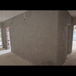 Kamil Hess - Ściana pokryta szarą, dekoracyjną gładzią w surowym wnętrzu. Widoczne drzwi w ościeżnicy i surowy beton na podłodze. Minimalistyczny efekt wykończenia.