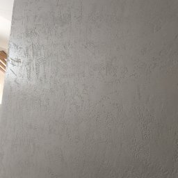 Kamil Hess - Ściana z szarą, dekoracyjną strukturą, imitującą efekt piasku. Widoczny fragment sufitu i oświetlenie. Tekstura ściany z bliska, z delikatnymi nierównościami.