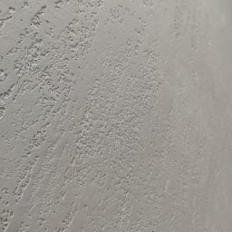 Kamil Hess - Szara ściana z dekoracyjną, chropowatą fakturą, wykonana techniką gładzi strukturalnej. Widoczne nieregularne wzory i delikatne cieniowanie. Ujęcie pod kątem.