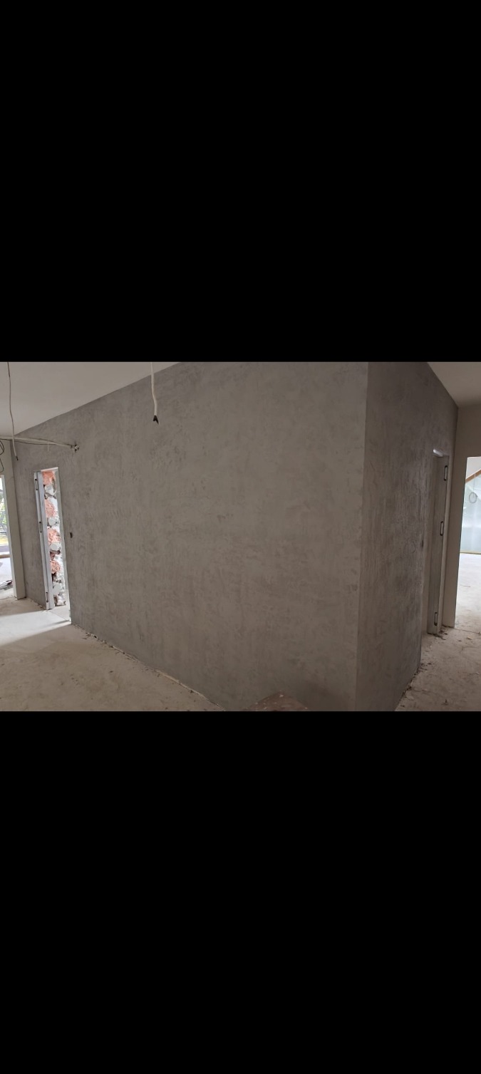 Ściana pokryta szarą, dekoracyjną gładzią w surowym wnętrzu. Widoczne drzwi w ościeżnicy i surowy beton na podłodze. Minimalistyczny efekt wykończenia.