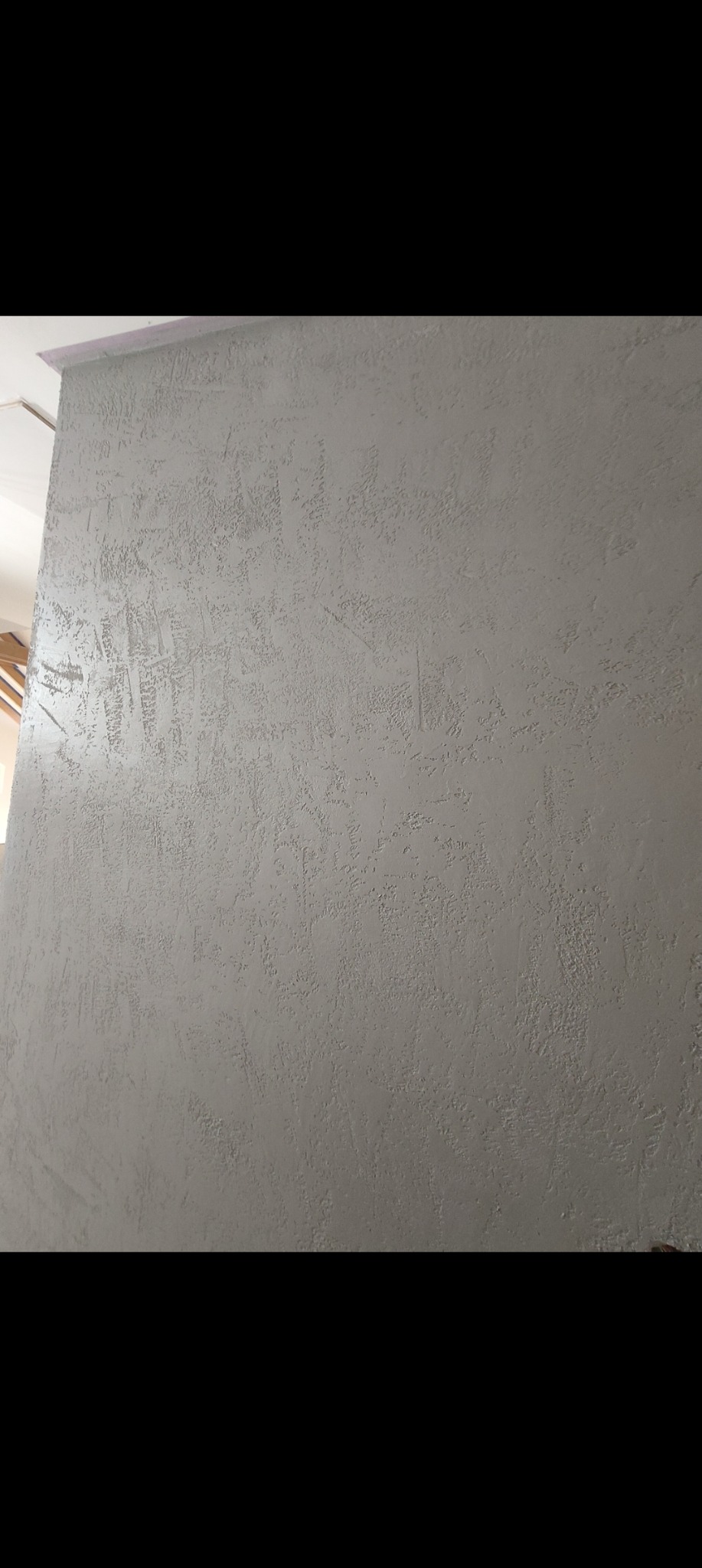 Ściana z szarą, dekoracyjną strukturą, imitującą efekt piasku. Widoczny fragment sufitu i oświetlenie. Tekstura ściany z bliska, z delikatnymi nierównościami.