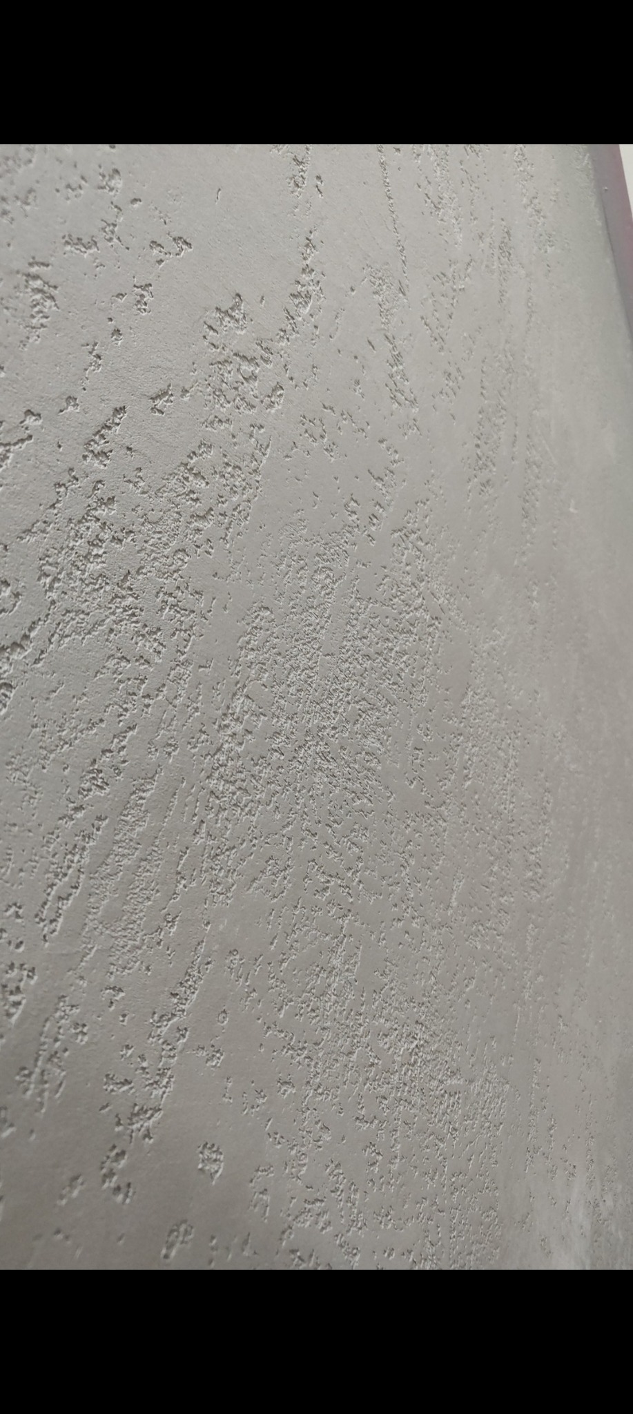 Szara ściana z dekoracyjną, chropowatą fakturą, wykonana techniką gładzi strukturalnej. Widoczne nieregularne wzory i delikatne cieniowanie. Ujęcie pod kątem.