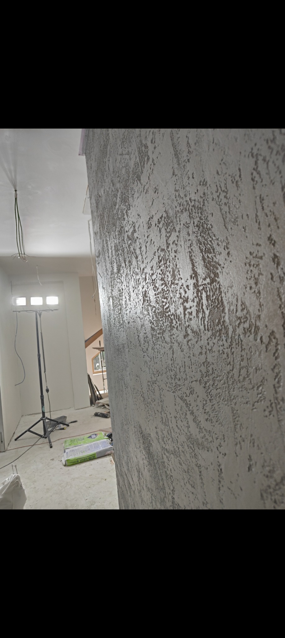 Ściana z dekoracyjną, szarą strukturą w nowym wnętrzu. Widoczny reflektor budowlany i worki z gładzią. Efektowna faktura ściany, podkreślona przez boczne oświetlenie.