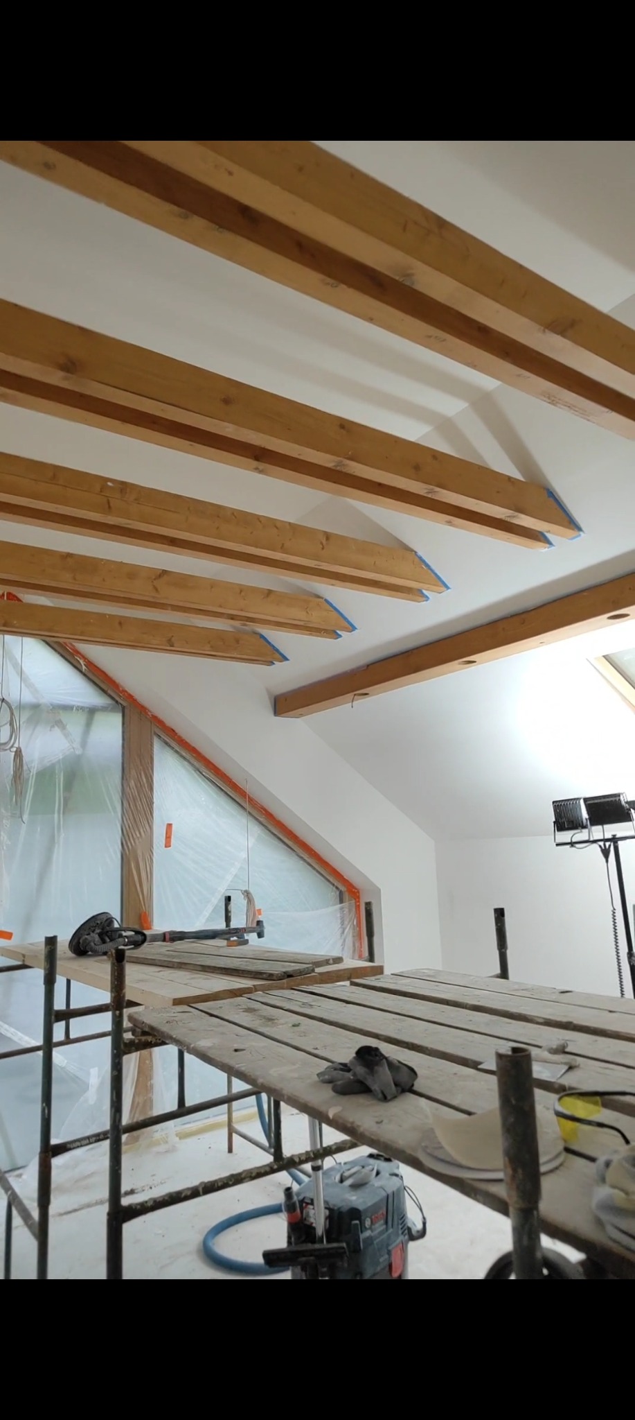 Skośny sufit z drewnianymi belkami zabezpieczonymi taśmą malarską, widok z rusztowania z odkurzaczem przemysłowym. Okno zabezpieczone folią malarską.