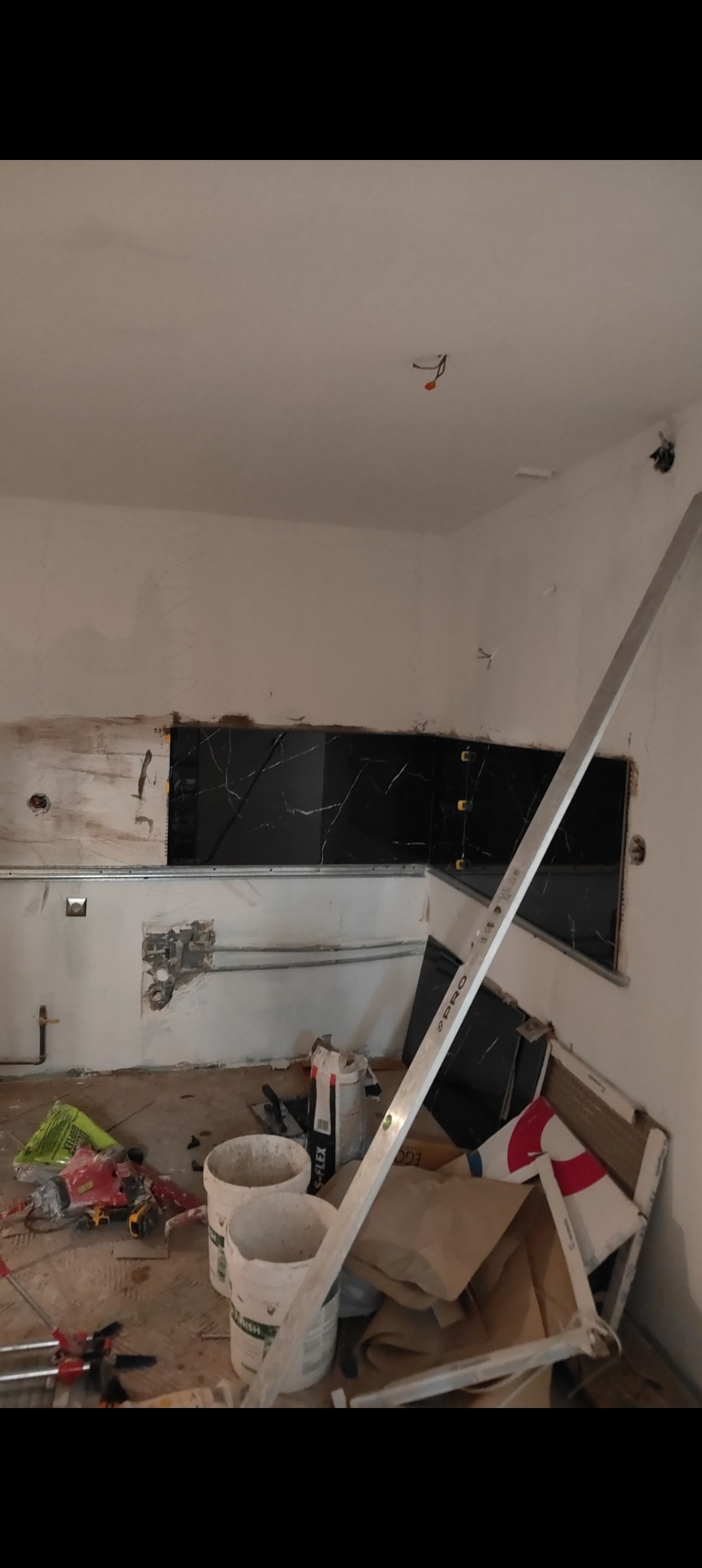 Częściowo wyłożona czarnymi płytkami ściana w kuchni podczas remontu. Widoczne narzędzia i materiały budowlane, poziomica oparta o ścianę.