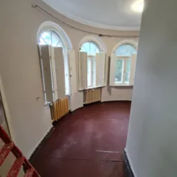 Wnętrze okrągłego pokoju po malowaniu ścian na jasny odcień, widoczne trzy okna z otwartymi okiennicami i czerwona podłoga. W rogu drabina malarska.