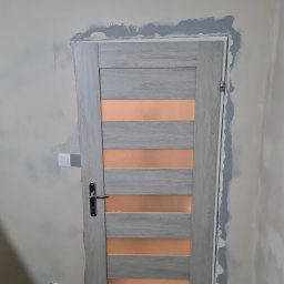 Kamil Rączak - Przygotowanie ściany wokół ościeżnicy do malowania. Widoczne ślady szpachlowania i gruntowania. Drzwi wewnętrzne z poziomymi przeszkleniami w kolorze drewna.