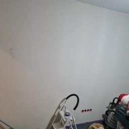 Kamil Rączak - Biała ściana w trakcie przygotowania do malowania, widoczne ślady po szpachlowaniu, drabina malarska i gniazdka elektryczne. Przygotowanie do prac wykończeniowych.