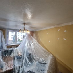 Kamil Rączak - Pokój w trakcie przygotowań do malowania: meble zabezpieczone folią, ściany z widocznymi punktowymi naprawami, sufit po nałożeniu gładzi, okno z widokiem na zewnątrz.