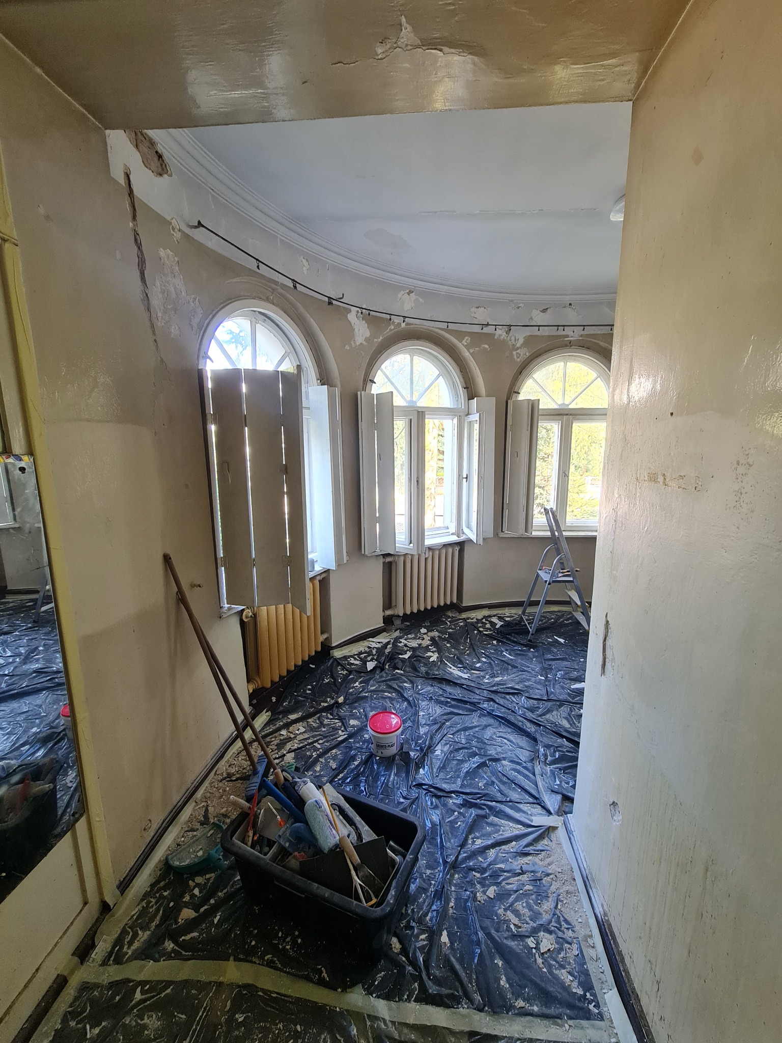 Pokój przed remontem: ściany z ubytkami, okna z otwartymi okiennicami, podłoga zabezpieczona folią, widoczne narzędzia i drabina. Przygotowanie do prac wykończeniowych.