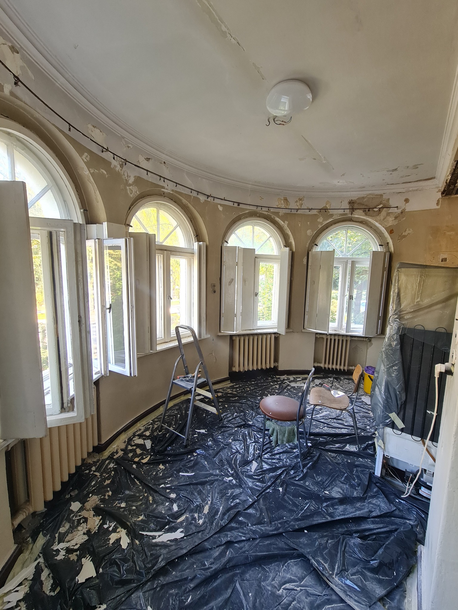 Okrągłe pomieszczenie w trakcie przygotowań do remontu: zabezpieczona folią podłoga, odsłonięte okna, ślady starej farby na ścianach i suficie, drabina i krzesła.