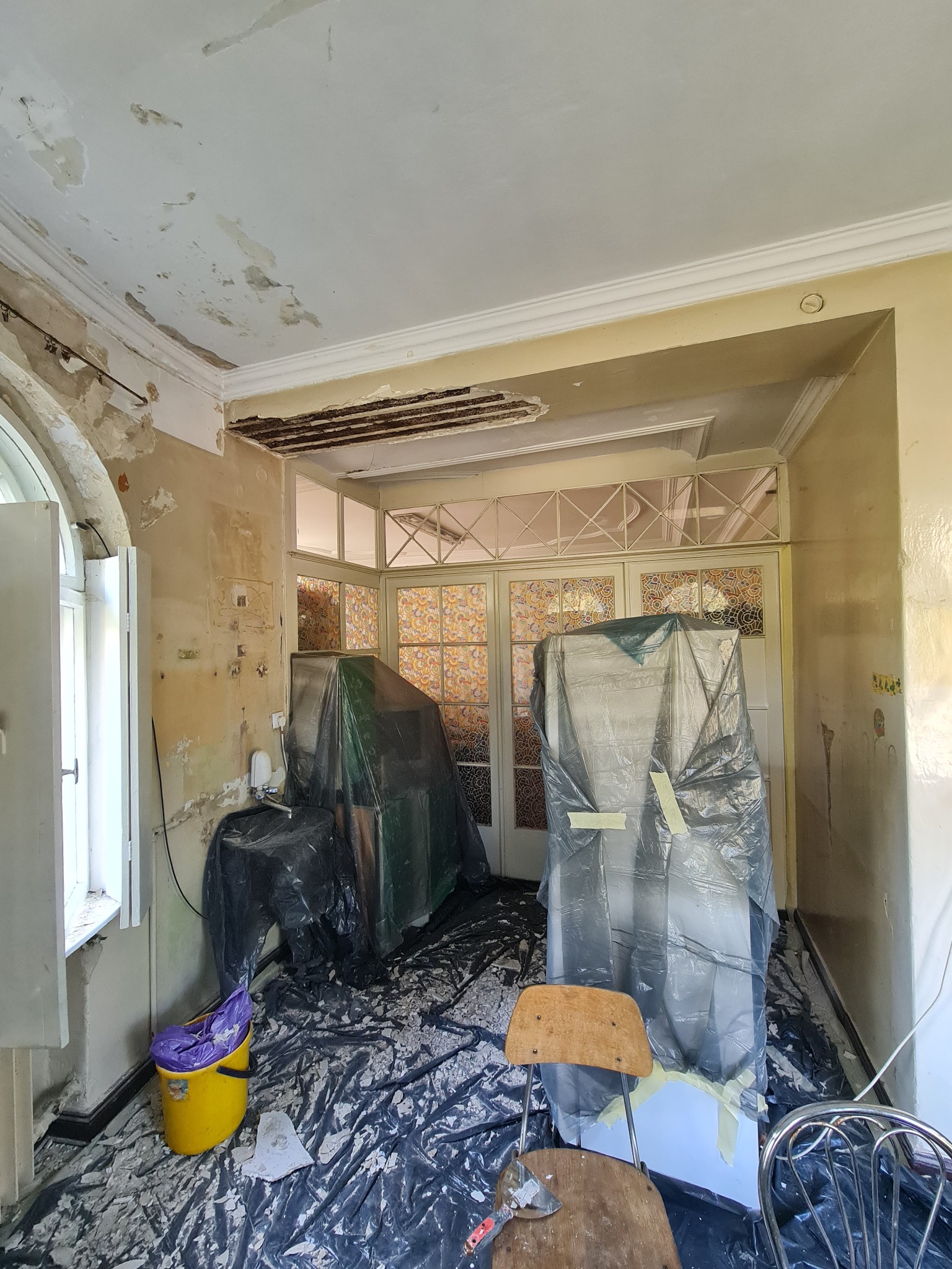 Wnętrze w trakcie remontu: odsłonięty strop, ściany z ubytkami tynku, zabezpieczone meble folią, wiadro i narzędzia na podłodze. Widoczne ślady prac przygotowawczych.