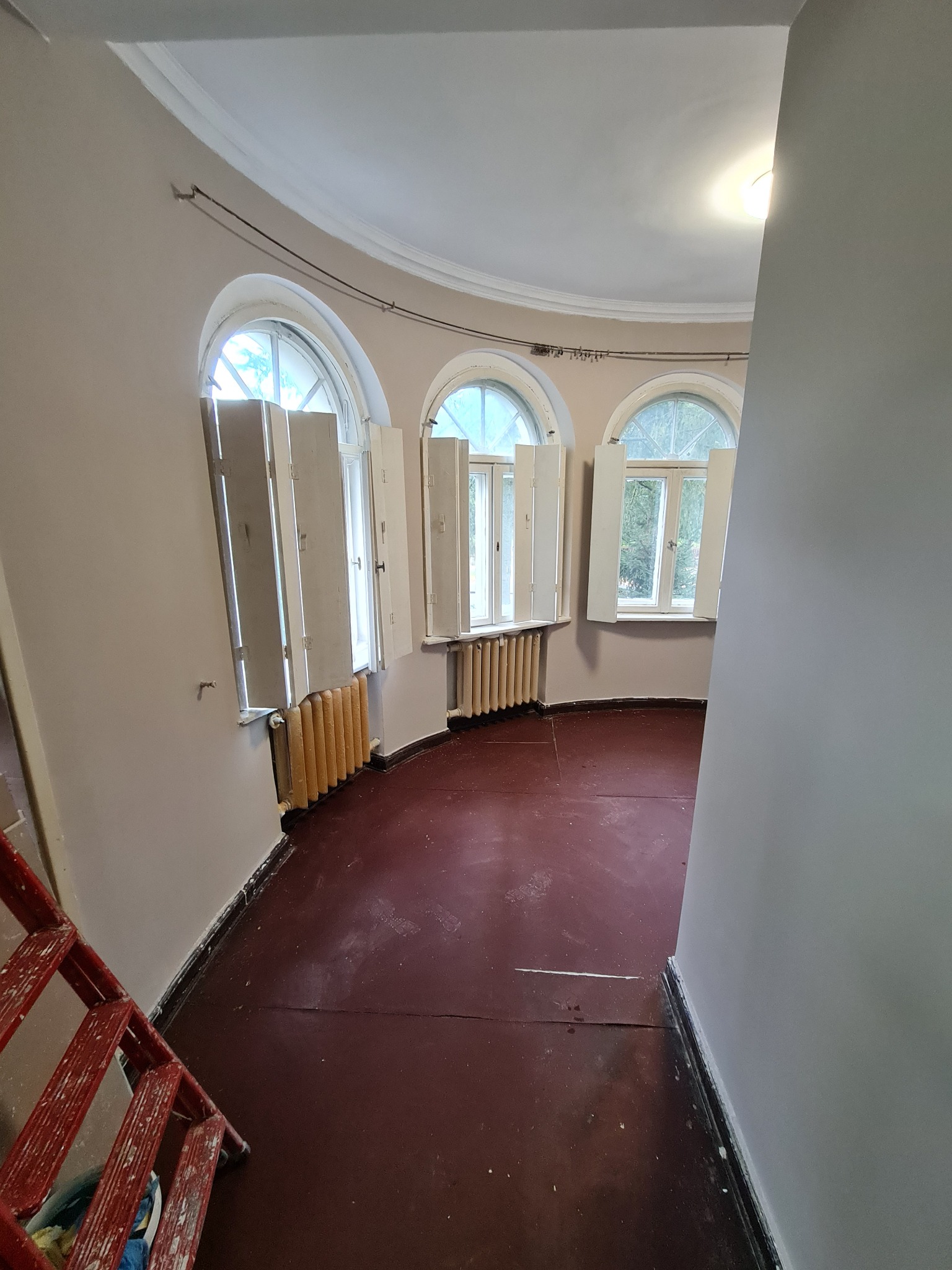 Wnętrze okrągłego pokoju po malowaniu ścian na jasny odcień, widoczne trzy okna z otwartymi okiennicami i czerwona podłoga. W rogu drabina malarska.