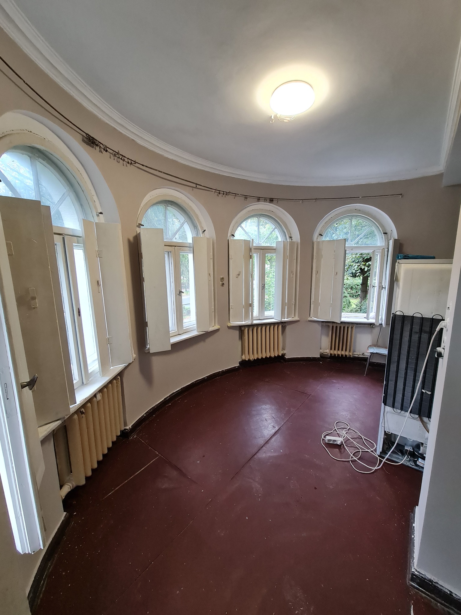 Okrągłe wnętrze z trzema łukowymi oknami z otwartymi okiennicami, brązową podłogą i grzejnikami. Widoczny fragment lodówki i przedłużacz na podłodze.