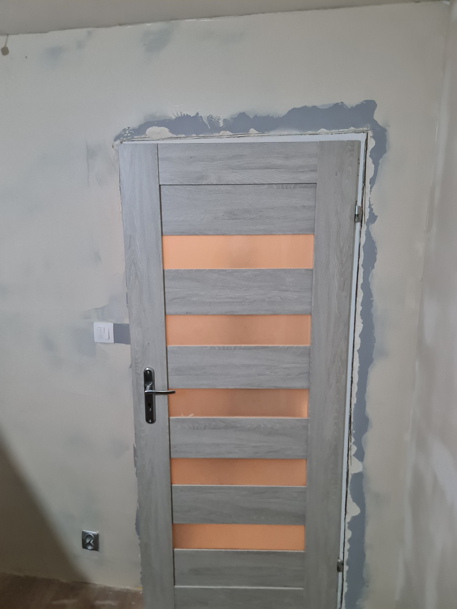 Przygotowanie ściany wokół ościeżnicy do malowania. Widoczne ślady szpachlowania i gruntowania. Drzwi wewnętrzne z poziomymi przeszkleniami w kolorze drewna.