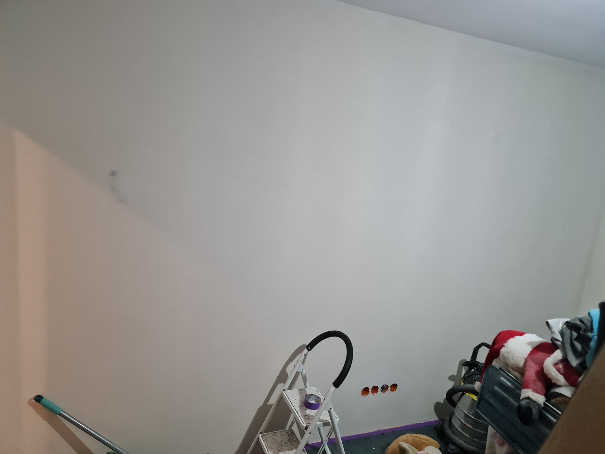 Biała ściana w trakcie przygotowania do malowania, widoczne ślady po szpachlowaniu, drabina malarska i gniazdka elektryczne. Przygotowanie do prac wykończeniowych.