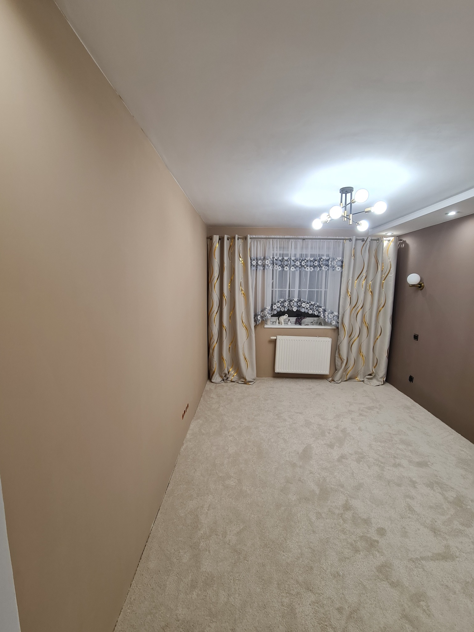 Beżowe ściany w pokoju po malowaniu, z kremowym dywanem i oknem z dekoracyjnymi zasłonami. Nowoczesna lampa sufitowa i grzejnik pod oknem.