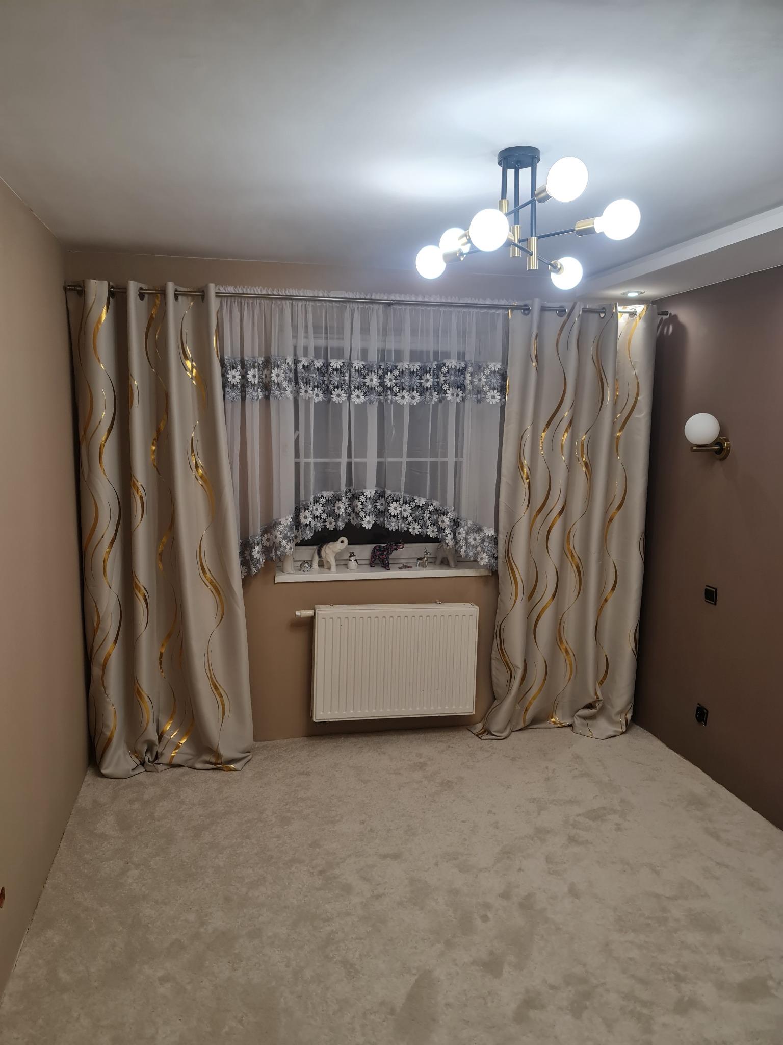 Jasne wnętrze po remoncie: beżowe ściany, dywan, dekoracyjne zasłony z motywem złotych fal i nowoczesna lampa sufitowa z kulistymi żarówkami.