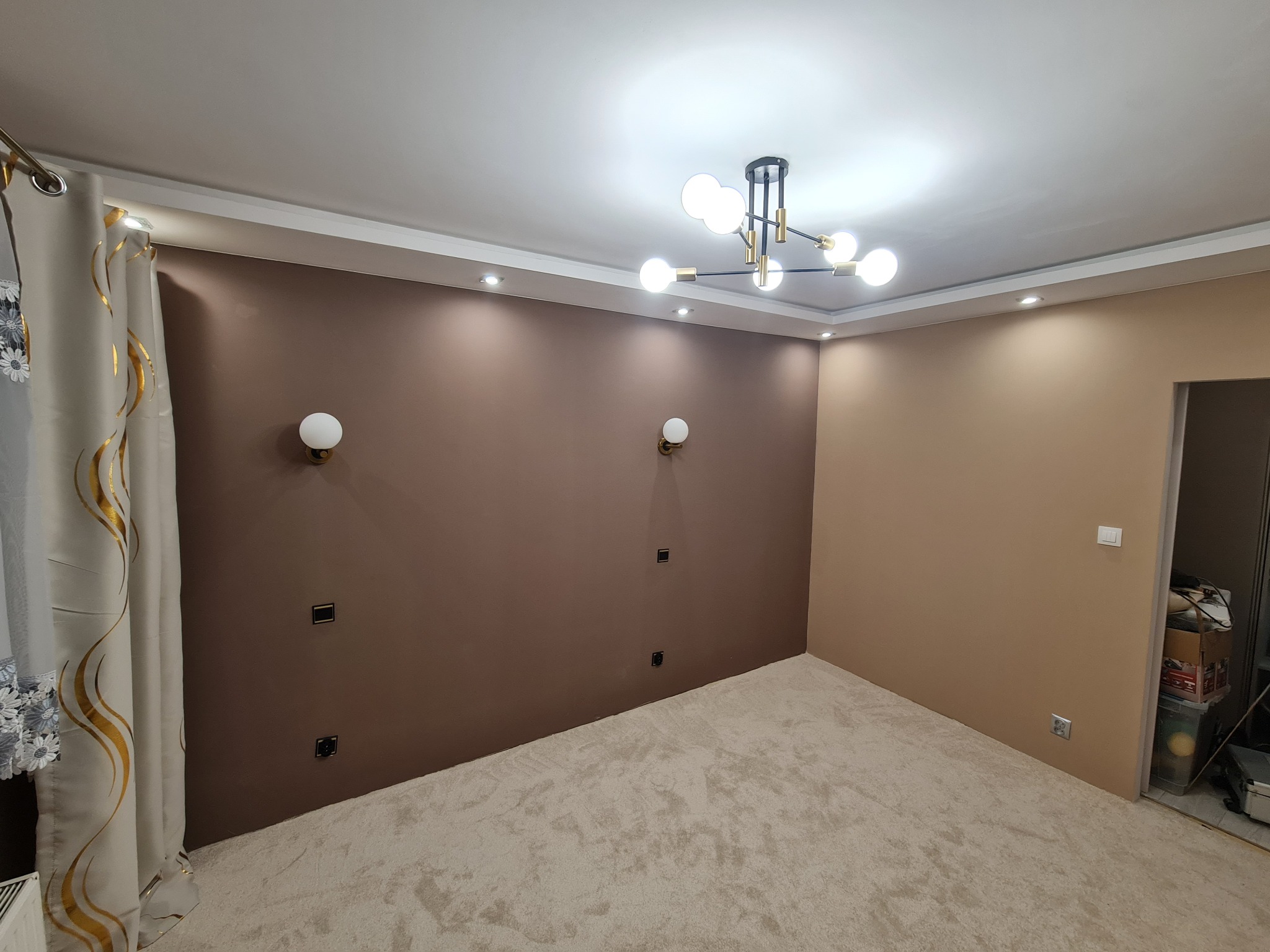 Pokój po malowaniu ścian na dwa odcienie brązu, z beżową wykładziną i nowoczesną lampą sufitową. Widoczne gniazdka elektryczne i dekoracyjne kinkiety.