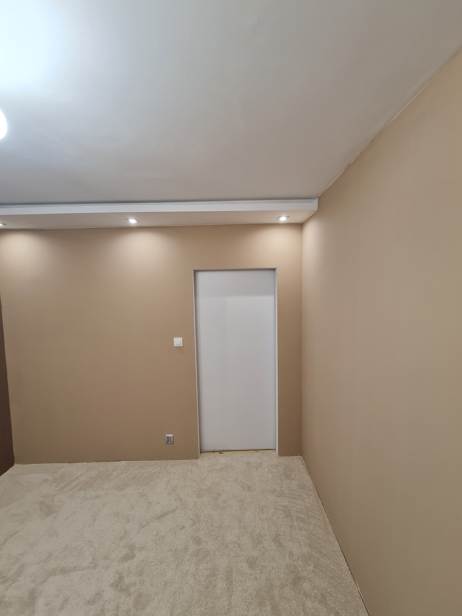Beżowe ściany w pokoju po malowaniu, z białymi drzwiami i jasną wykładziną. Oświetlenie sufitowe punktowe. Widoczny fragment podłogi i gniazdko elektryczne.