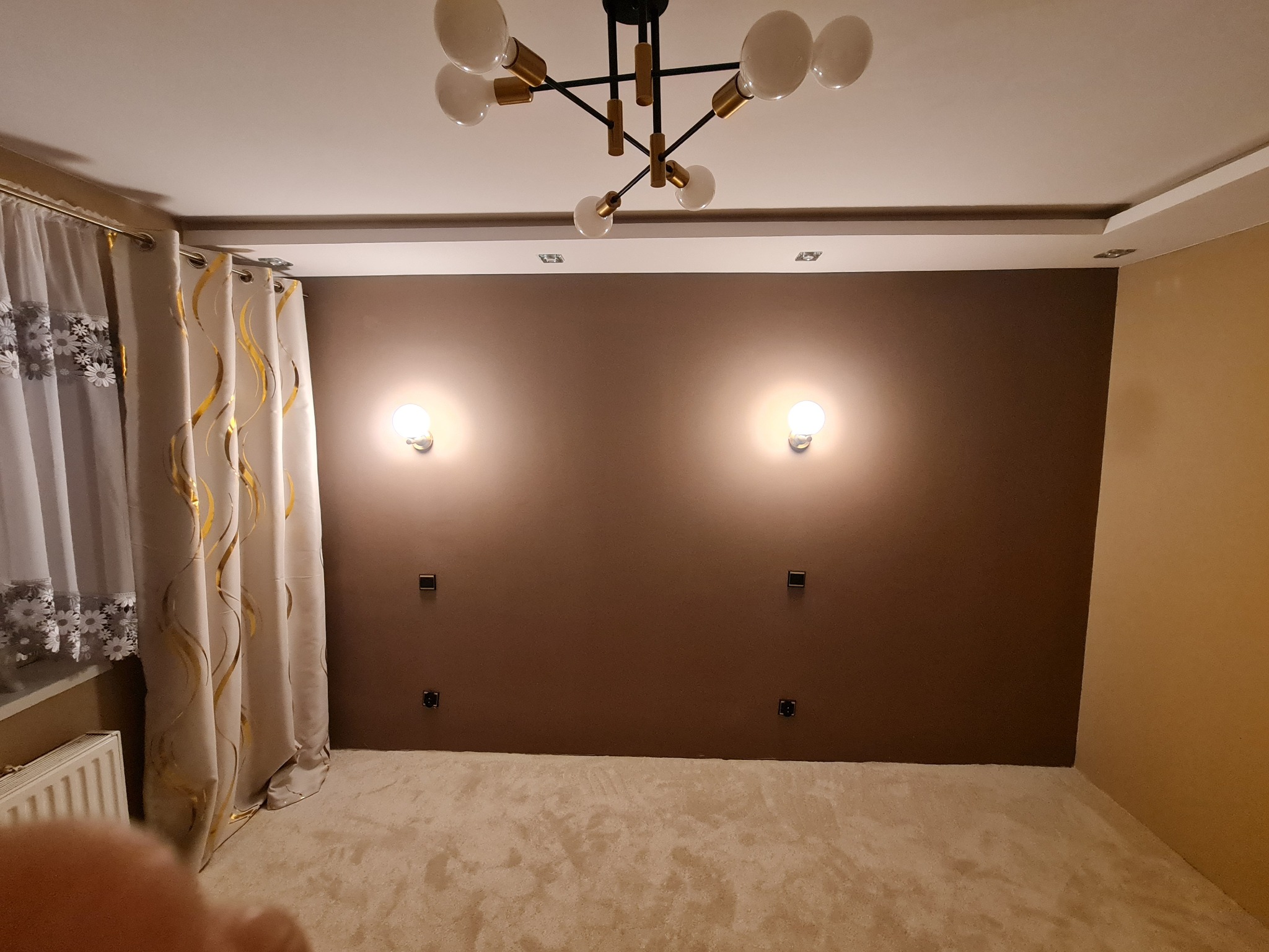 Pokój po malowaniu ścian na brązowo, z kremową wykładziną i nowoczesną lampą sufitową. Widoczne gniazdka elektryczne i zasłony w oknie.