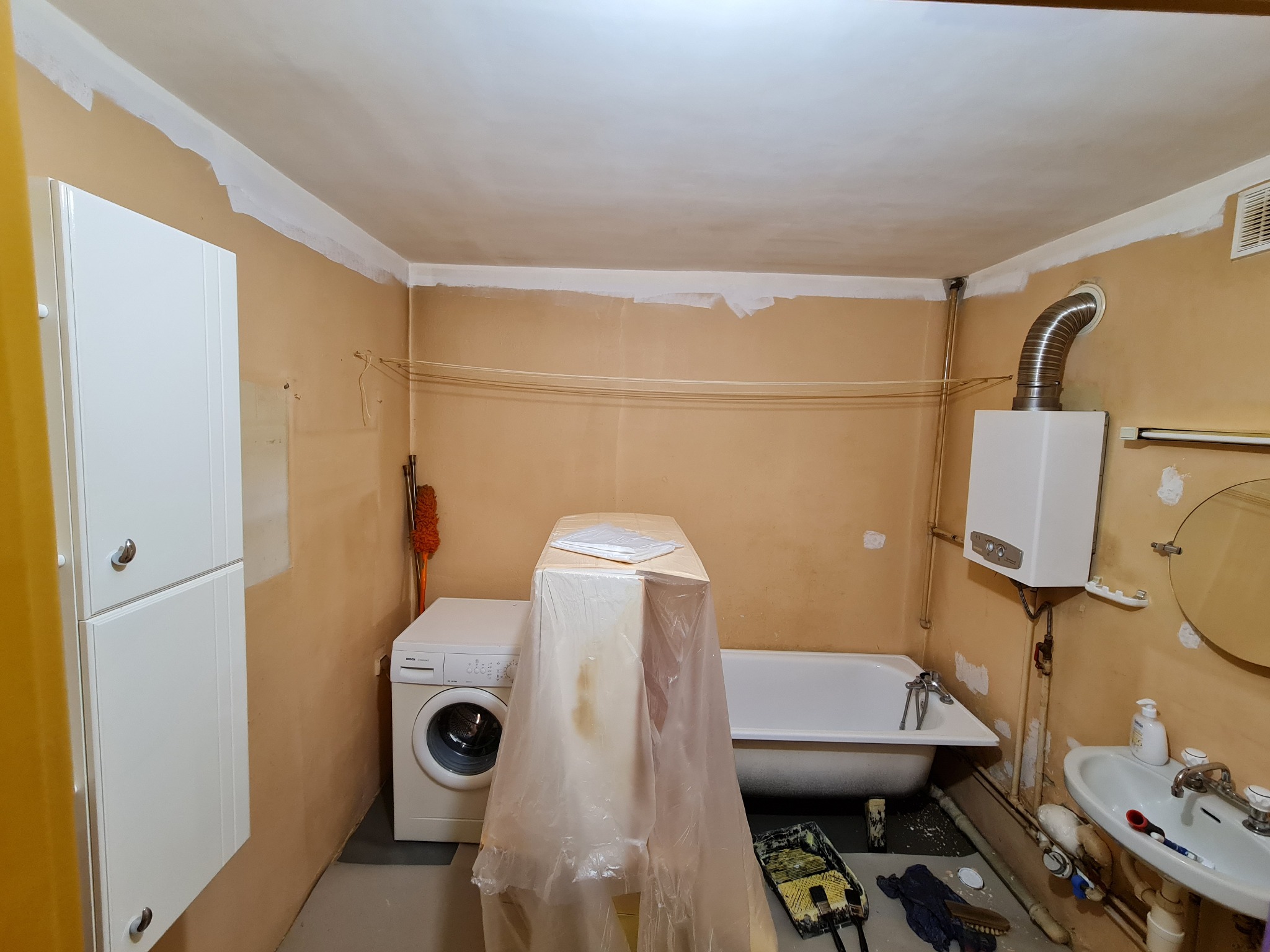 Remont łazienki w trakcie: ściany przygotowane do malowania, zabezpieczona pralka i wanna folią malarską, widoczne narzędzia malarskie. Świeżo zagruntowane łączenia ścian i sufitu.