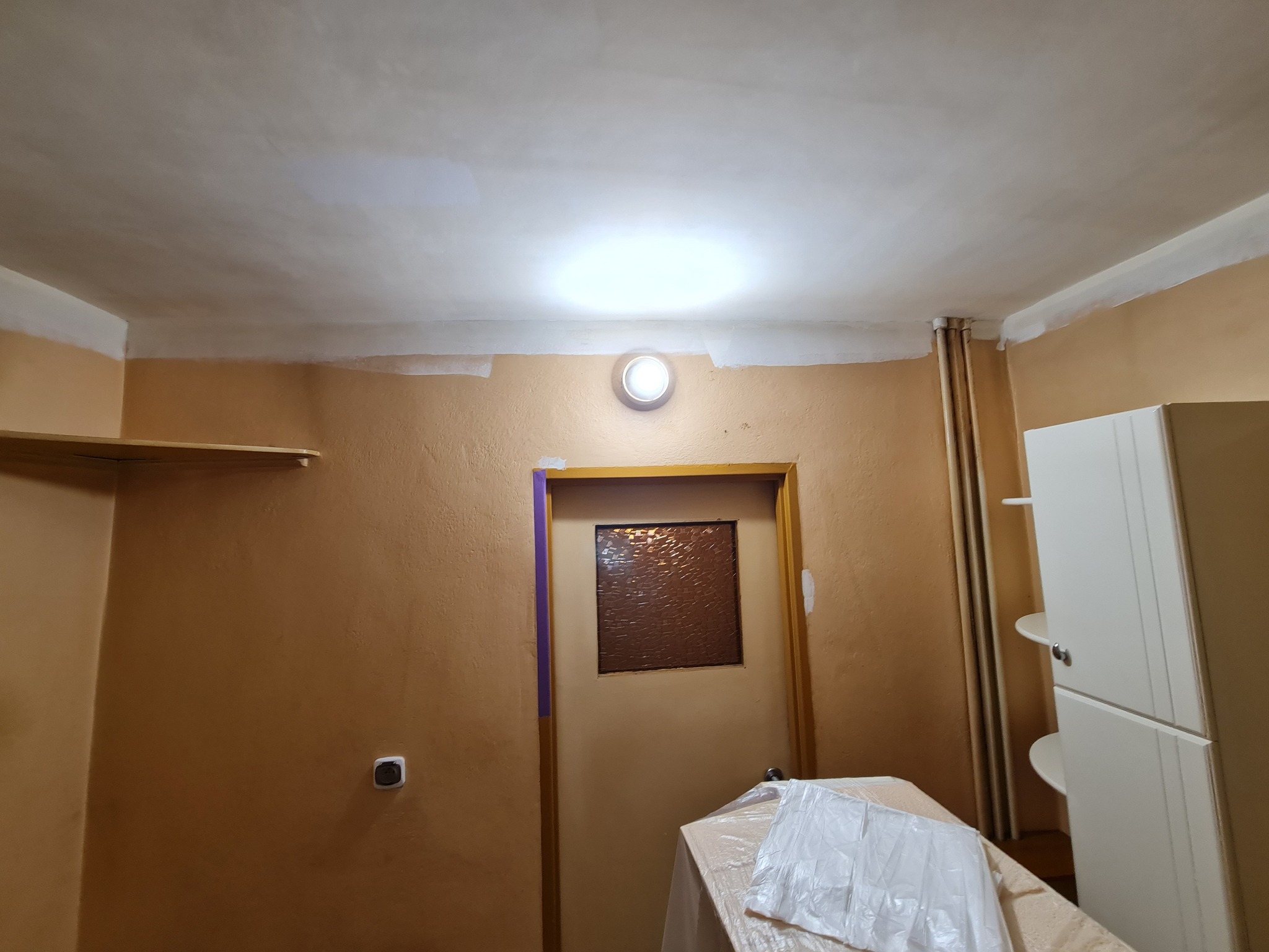 Pokój w trakcie malowania: świeżo pomalowany sufit, ściany w odcieniach beżu, zabezpieczone listwy i futryny. Widoczne przygotowania do dalszych prac.