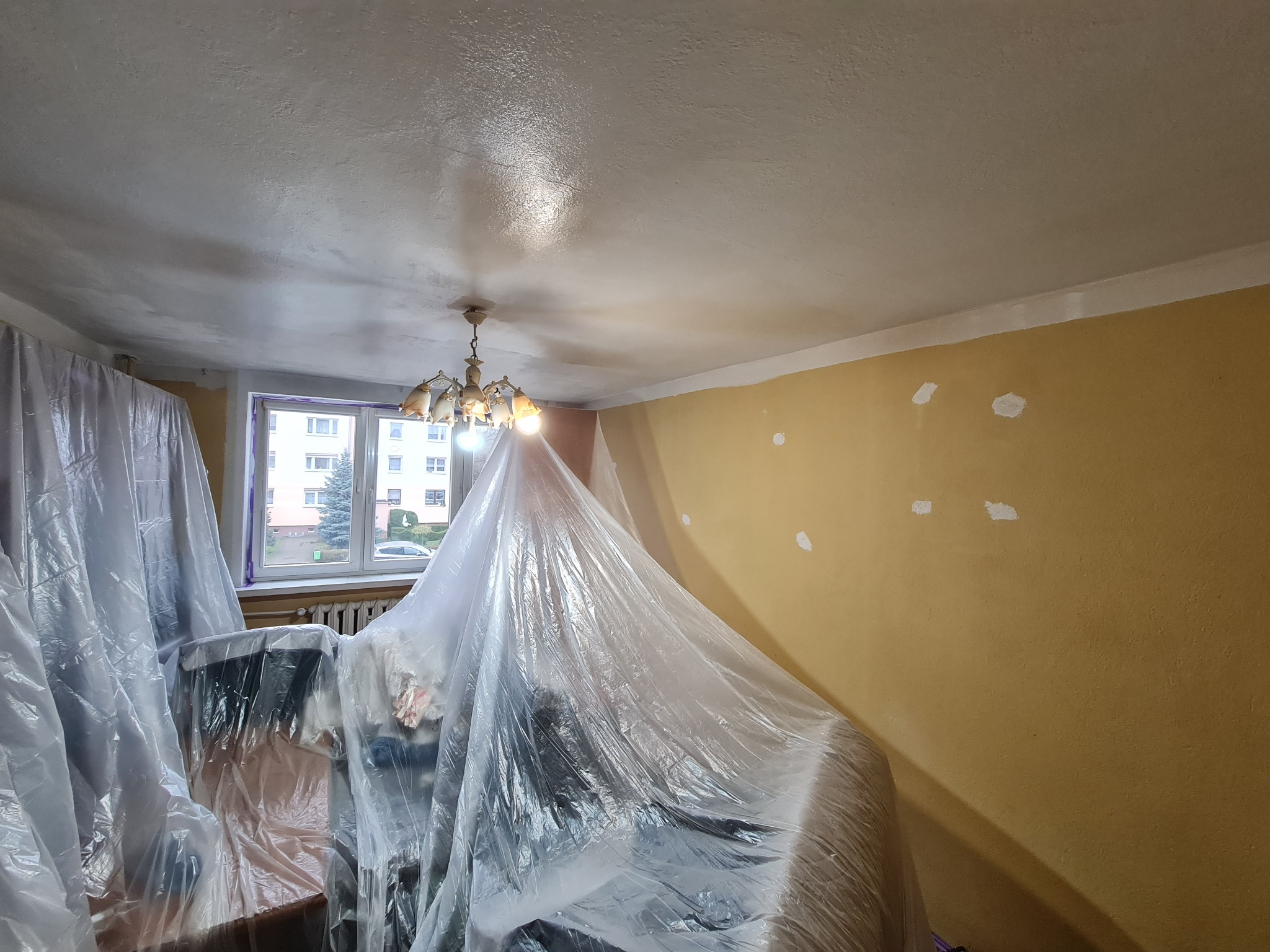 Pokój w trakcie przygotowań do malowania: meble zabezpieczone folią, ściany z widocznymi punktowymi naprawami, sufit po nałożeniu gładzi, okno z widokiem na zewnątrz.