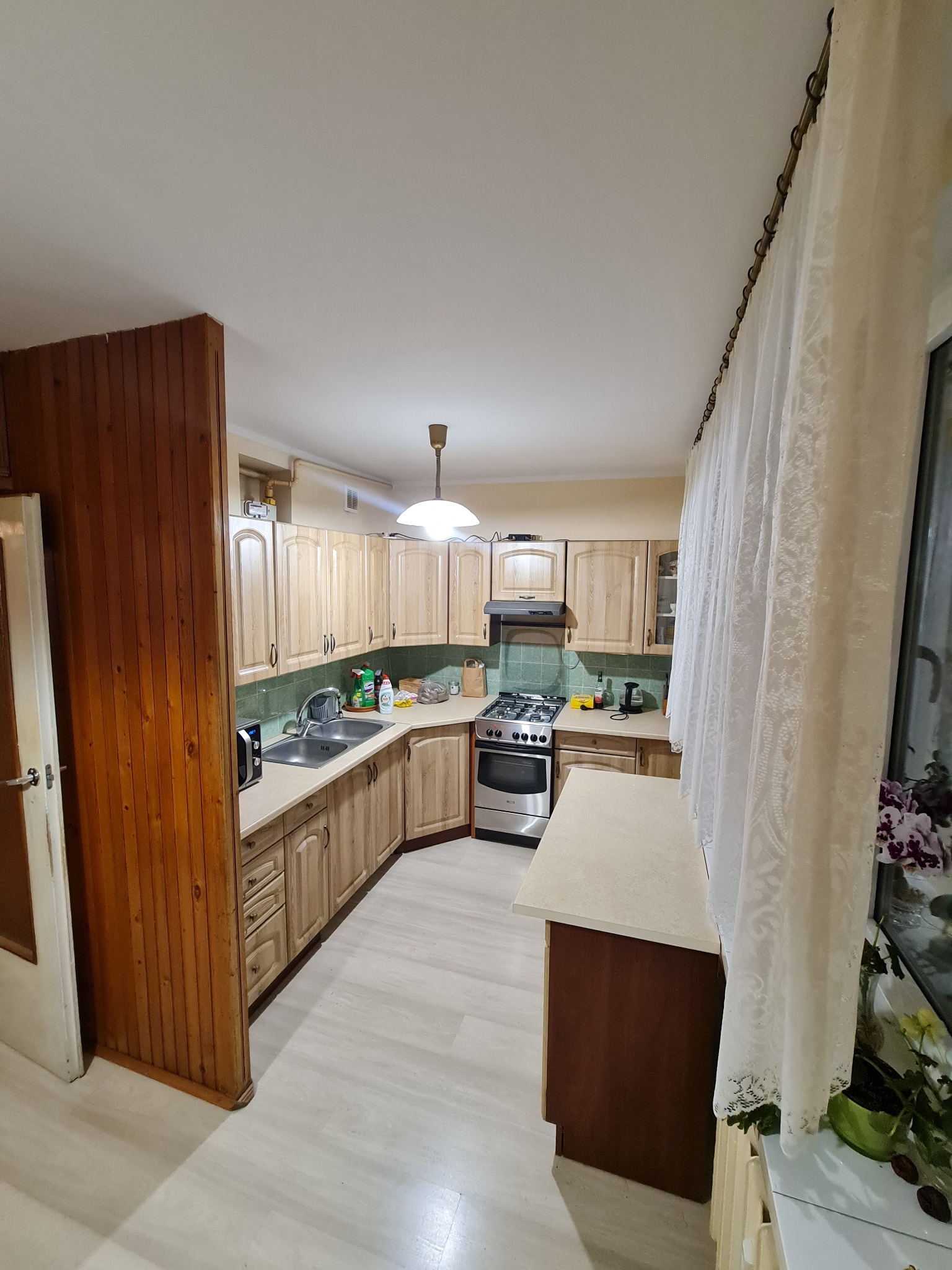 Jasna kuchnia z drewnianymi szafkami, zielonymi płytkami nad blatem i białymi zasłonami w oknie. Widok na kuchenkę i zlew. Całość utrzymana w ciepłej tonacji.