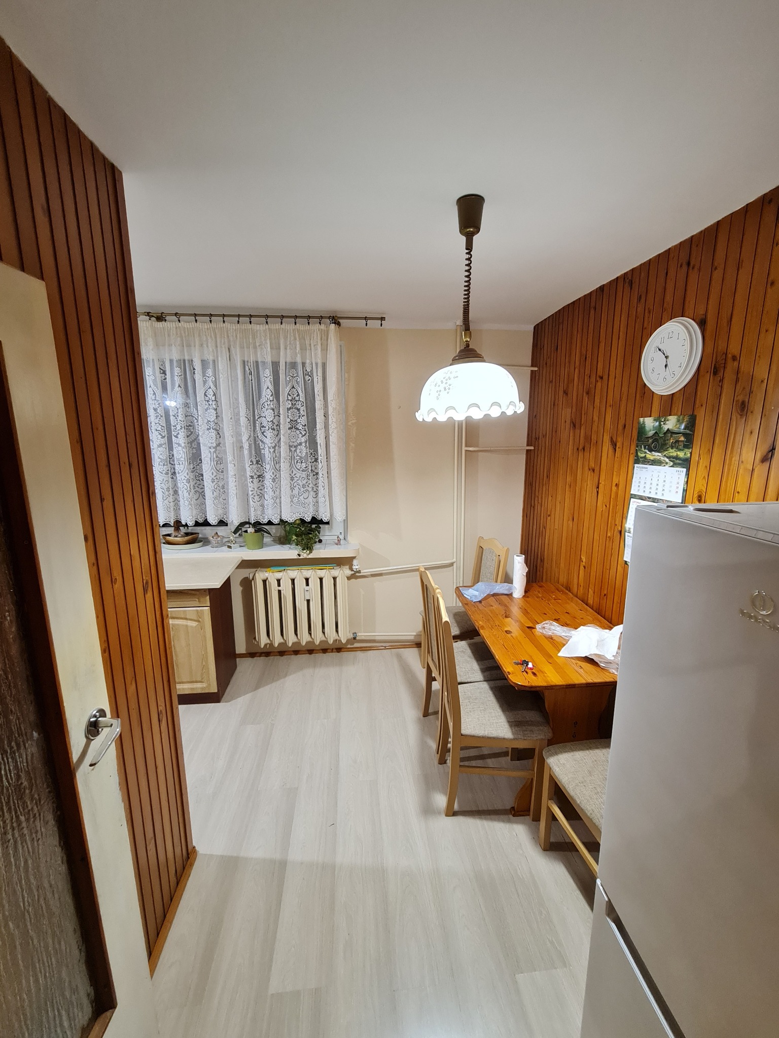 Jasna kuchnia z drewnianą boazerią, stołem i krzesłami. Widok na okno z firanką i grzejnik. Podłoga wyłożona panelami. W tle lodówka i wisząca lampa.