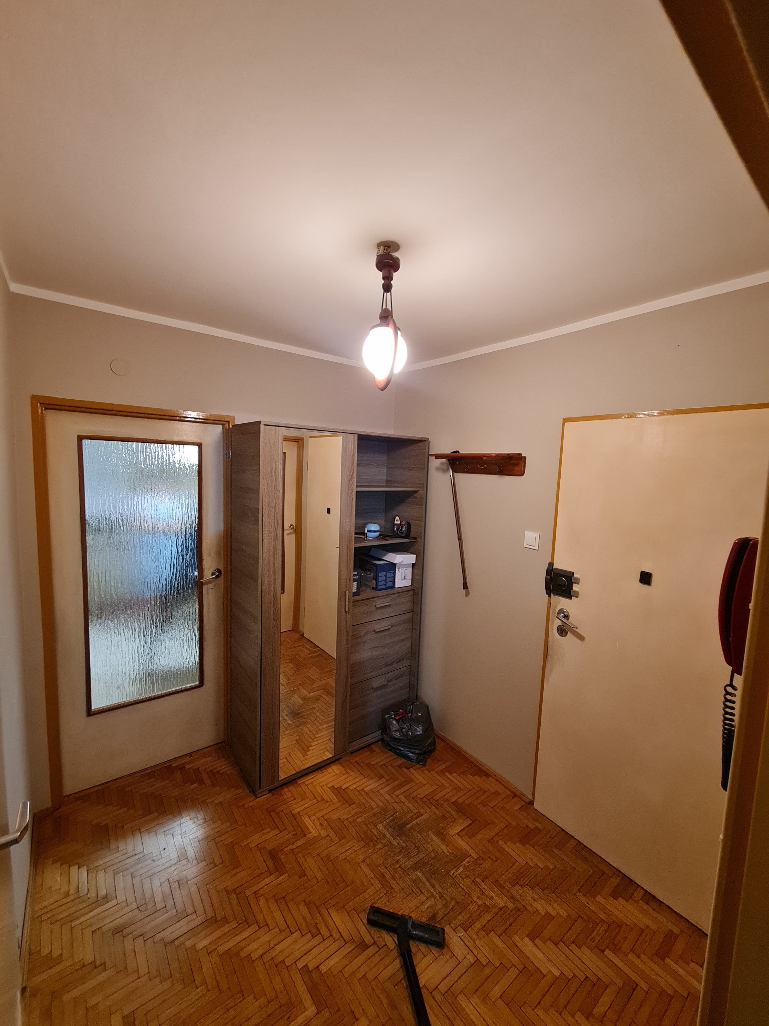 Pokój z parkietem w jodełkę, szafą z lustrem i odkurzaczem. Ściany w odcieniach beżu, sufit biały z lampą. Widoczne drzwi i elementy wyposażenia.