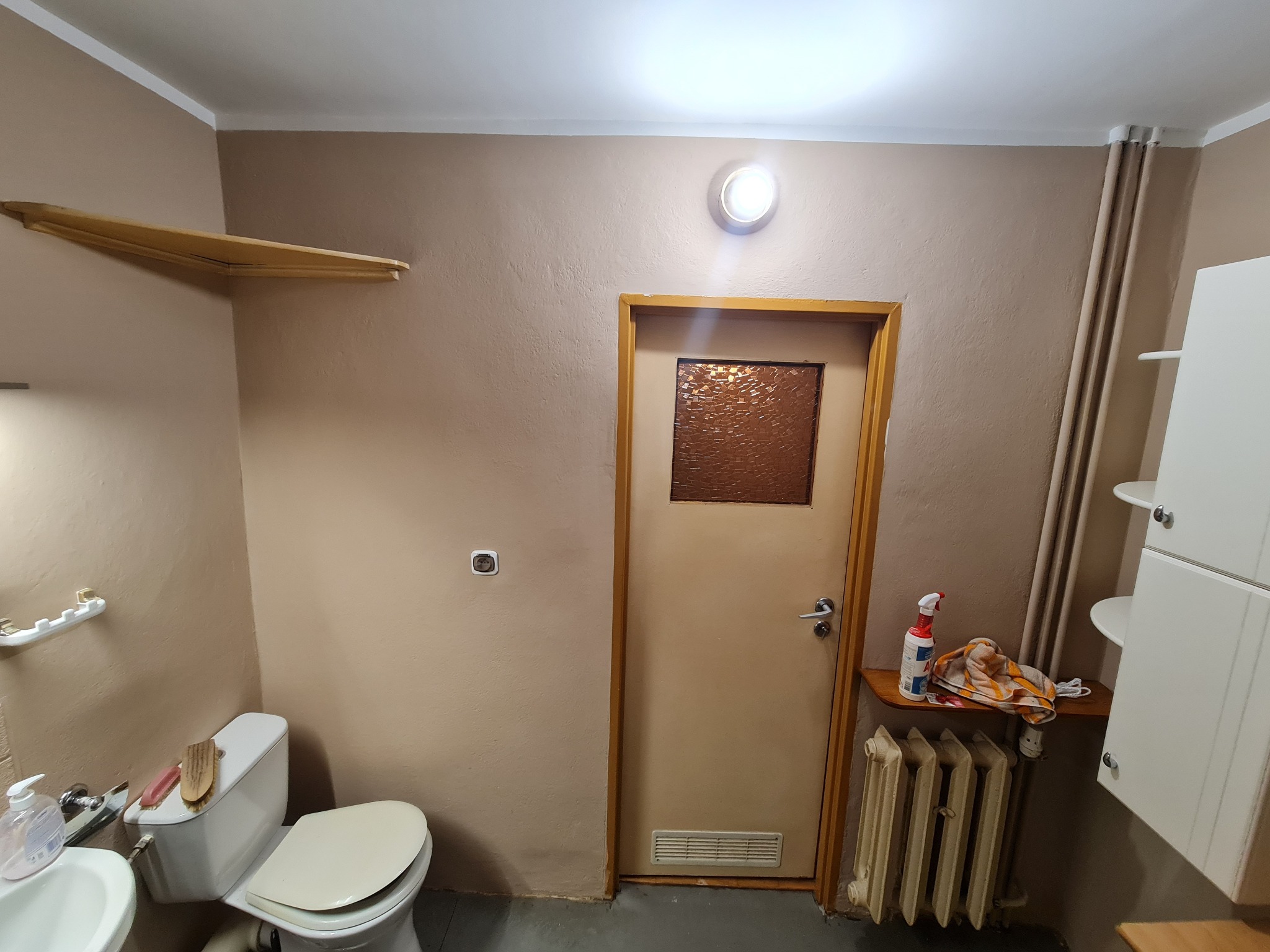 Beżowa łazienka po odświeżeniu: ściany w odcieniach beżu, biała armatura, grzejnik i drewniane półki. Widoczne detale, takie jak drzwi z szybą i oświetlenie.