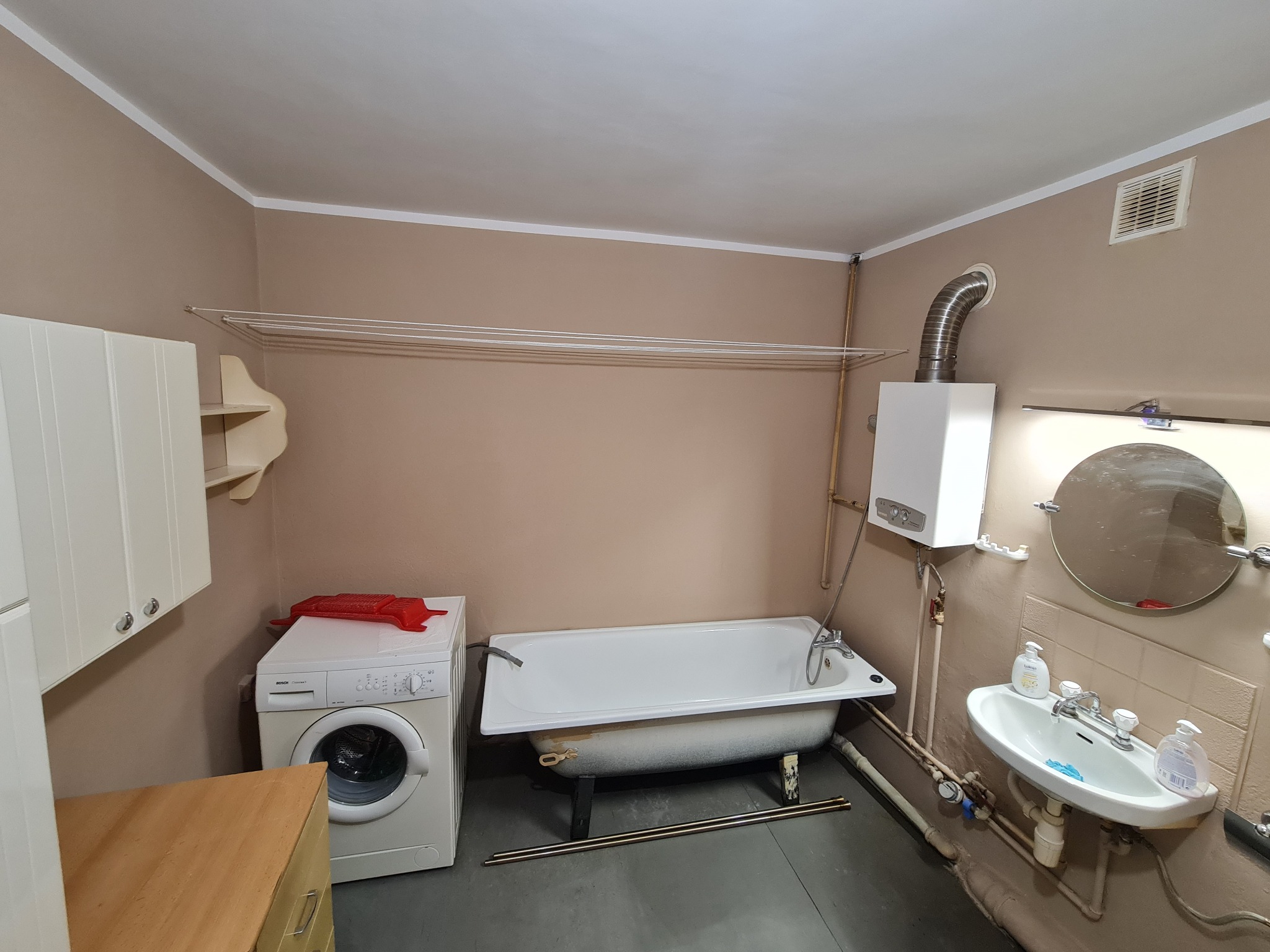 Wyremontowana łazienka w beżowych odcieniach z wanną, pralką i umywalką. Widoczny bojler i suszarka podsufitowa. Minimalistyczny styl, funkcjonalne rozwiązanie.