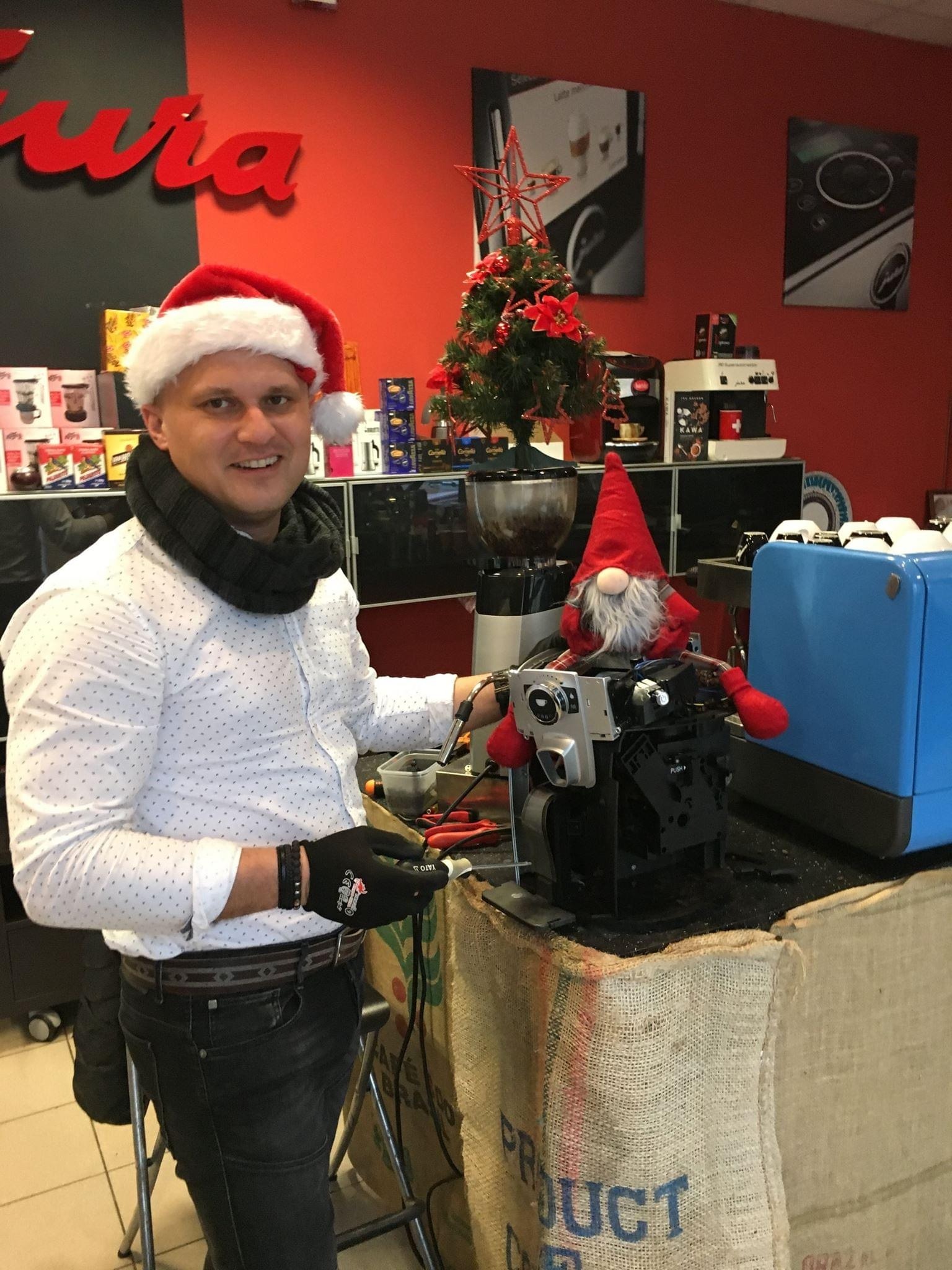 Serwisant w czapce Mikołaja naprawia ekspres do kawy Jura, obok stoi świąteczny skrzat. W tle ekspresy i dekoracje świąteczne w punkcie serwisowym.