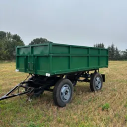 Zielona przyczepa rolnicza na polu po żniwach, widok z boku, szare opony, czarna rama, pochmurne niebo w tle, drzewa w oddali.