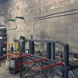 Wiktor Gronowski Usługi Ślusarskie - Metalowa konstrukcja w warsztacie, prawdopodobnie brama wjazdowa w trakcie spawania. Widoczny spawacz w tle, narzędzia i ceglana ściana.