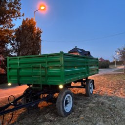Wiktor Gronowski Usługi Ślusarskie - Zielona przyczepa rolnicza z oświetleniem, stojąca na trawie o zmierzchu. Widoczne detale konstrukcyjne i oświetlenie, tło z domami i latarnią.