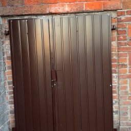 Wiktor Gronowski Usługi Ślusarskie - Brązowa, dwuskrzydłowa brama z metalu, zamontowana w murze z czerwonej cegły. Widoczne mocowania i zamek. Brama z pionowymi panelami.