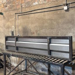 Wiktor Gronowski Usługi Ślusarskie - Metalowa konstrukcja bramy wjazdowej w warsztacie, na stole spawalniczym. Surowy stan, widoczne spawy i elementy konstrukcyjne. Tło to ceglana ściana i warsztatowy nieład.