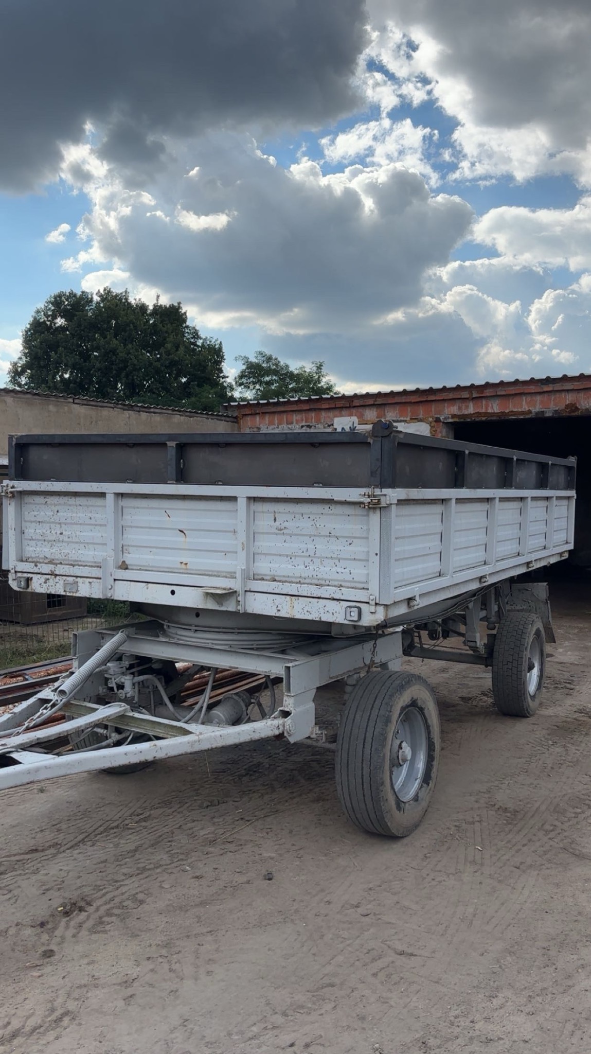 Przyczepa rolnicza z metalową konstrukcją, widoczna na tle budynku i pochmurnego nieba. Konstrukcja spawana, pomalowana na szaro.