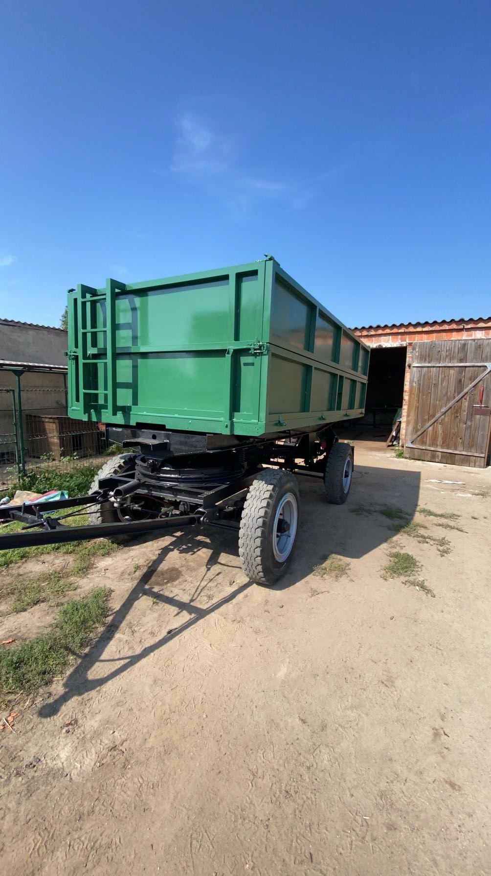 Zielona przyczepa rolnicza z drabinką, stojąca na piaszczystym podłożu przed budynkiem gospodarczym z otwartymi drewnianymi wrotami, w słoneczny dzień.