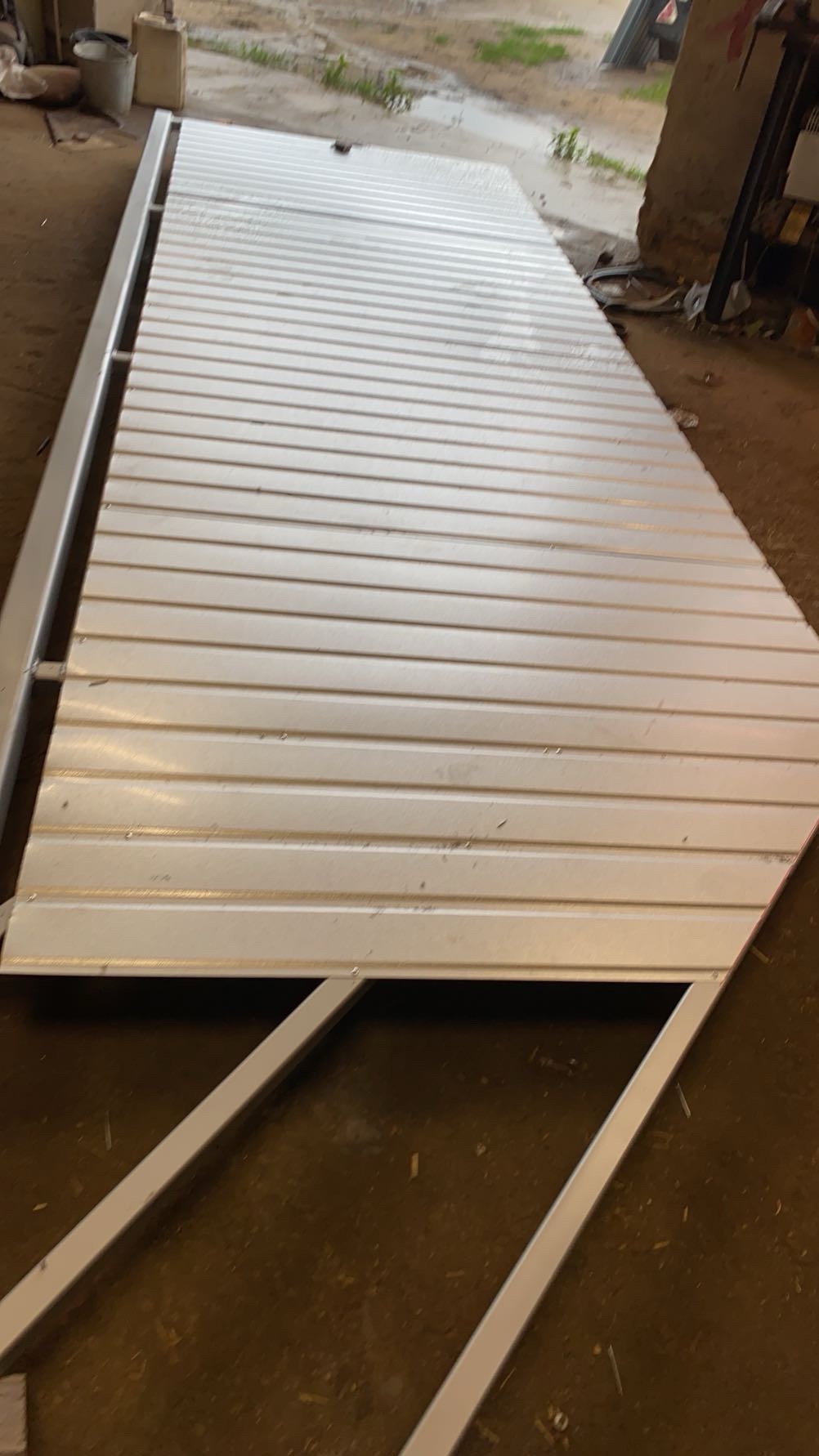 Biała, metalowa konstrukcja z poziomymi panelami, prawdopodobnie element bramy, leżąca na podłodze w warsztacie. Widoczne spawane łączenia i surowe wykończenie.