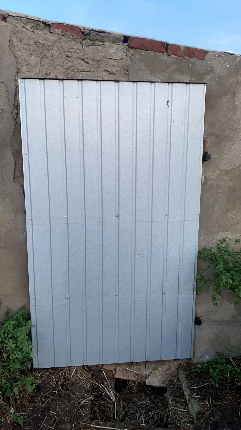 Metalowe, srebrne zabezpieczenie otworu w starej, betonowo-ceglanej ścianie. Widoczne ślady napraw i roślinność u podstawy. Proste, funkcjonalne rozwiązanie.