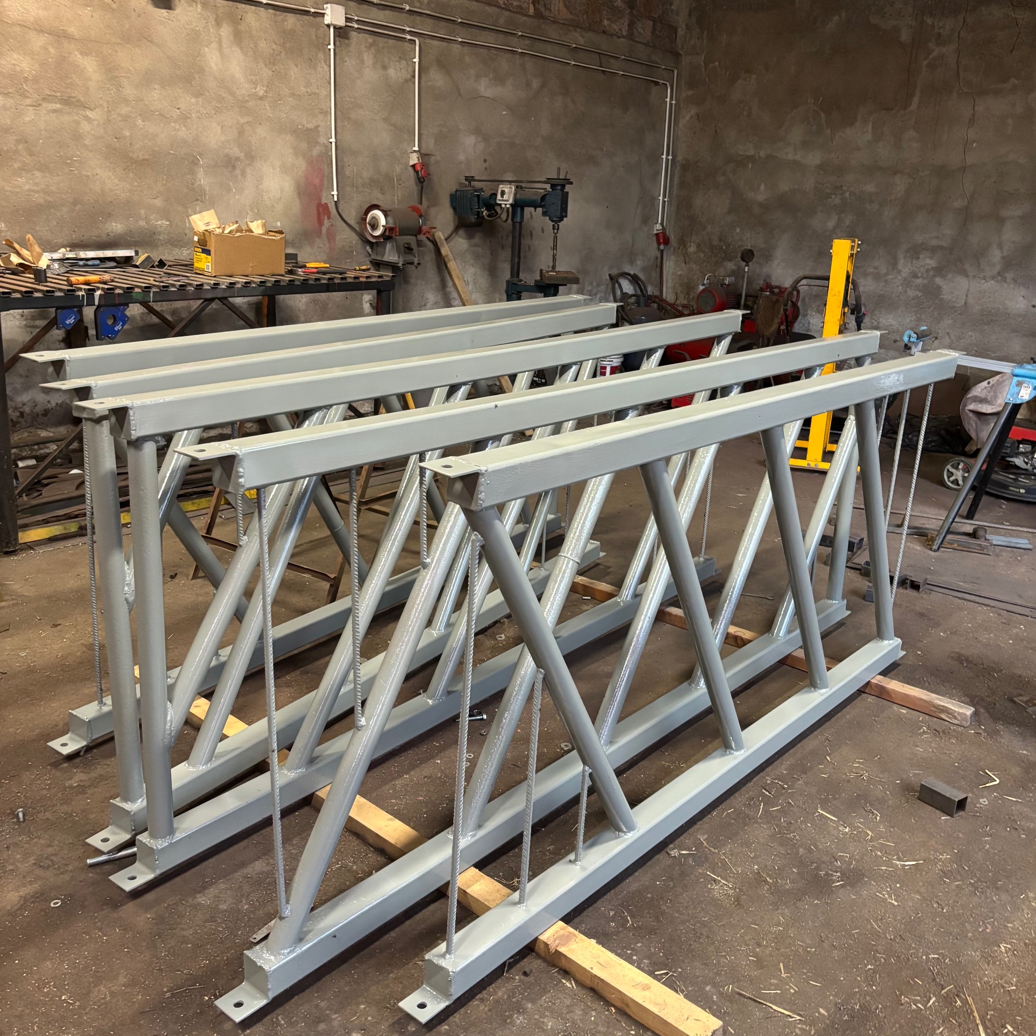Gotowe, pomalowane na szaro konstrukcje metalowe z profili, leżące w warsztacie. Widoczne narzędzia i maszyny ślusarskie w tle. Charakterystyczne spawy i mocowania.