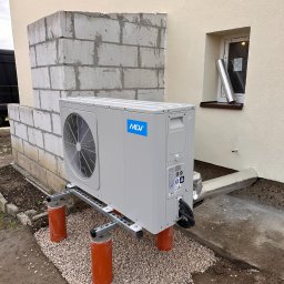 AQUAPULS SPÓŁKA Z OGRANICZONĄ ODPOWIEDZIALNOŚCIĄ - Szara jednostka zewnętrzna klimatyzacji MDV na pomarańczowych wspornikach, z widocznymi przewodami i fragmentem elewacji budynku w tle.