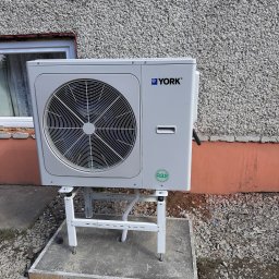 AQUAPULS SPÓŁKA Z OGRANICZONĄ ODPOWIEDZIALNOŚCIĄ - Biała jednostka zewnętrzna klimatyzacji York na betonowym fundamencie, zamontowana przy ścianie budynku o szarej, chropowatej fakturze. Widoczny zielony symbol R32.