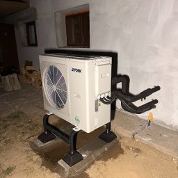 AQUAPULS SPÓŁKA Z OGRANICZONĄ ODPOWIEDZIALNOŚCIĄ - Biała pompa ciepła York na czarnej podstawie, z widocznymi rurami, zainstalowana przy szarej ścianie budynku z małymi oknami. Widoczne ślady po montażu.